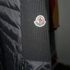 Svart pufferjacka från Moncler - Svart pufferjacka från Moncler med stickade ärmar och huva. Jackan har quiltad front i syntetmaterial, dragkedja och Moncler-logga på ärmen. Snygg kombination av stickad och puffer, perfekt för kyliga dagar.