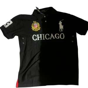 Svart Polo Ralph Lauren Chicago piké - Svart pikétröja från Polo Ralph Lauren med broderad gul och röd logga, stor vit text 'CHICAGO' och ikonisk polospelare på bröstet. Nummer 3 på ryggen och ärmen. Slim fit-modell med klassisk krage och knappar framtill.