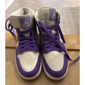 Nike Air Jordan 1 mid lila/vita sneakers - Nike Air Jordan 1 sneakers i lila och vitt med klassisk hög siluett. Skorna har lila snörning, perforerad tå och ikoniska Jumpman-loggan på plösen. Ovandelen är i skinn och textil, med vit bas och lila detaljer. Perfekta för dig som vill sticka ut med en färgstark look. Använda en gång
