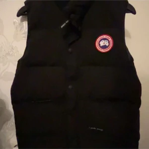 Svart dunväst från Canada Goose - Svart dunväst från Canada Goose med klassisk logga på bröstet. Västen är quiltad och har knappar framtill. Perfekt för lager-på-lager och håller dig varm under kyliga dagar.