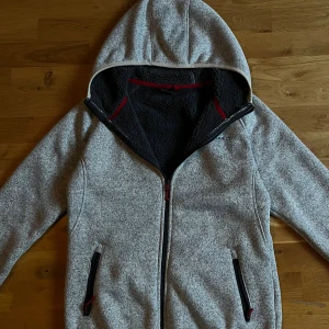 Grå teddyjacka med huva från Bluewear - Mysig gråmelerad teddyjacka från Bluewear med svart fluffigt foder, huva och hel dragkedja. Jackan har två fickor med svarta dragkedjor och röda detaljer. Perfekt för kalla dagar när du vill vara både varm och chill.