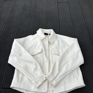 Vit jacka från Vero Moda - Vit jacka från Vero Moda med krage, tryckknappar och två stora bröstfickor. Jackan har en rak passform. 