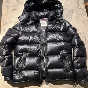 Svart Moncler pufferjacka med huva - Svart pufferjacka från Moncler med glansig finish, stor huva och två dragkedjeförsedda fickor. Jackan har klassisk Moncler-logga på ärmen och breda, quiltade sektioner. Perfekt för kalla dagar och har en riktigt fet streetstil.