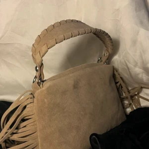 Beige mocka handväska med fransar - Snygg beige handväska i mocka med flätat handtag och coola fransdetaljer på sidorna. Väskan har en enkel, rektangulär form och metallringar som fäster handtaget. Perfekt för dig som gillar boho-vibe och vill sticka ut med detaljer.