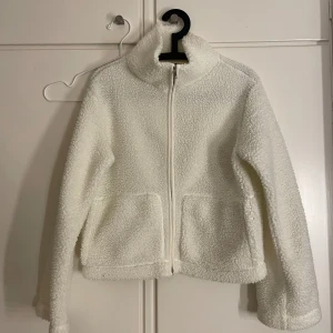 Vit teddyjacka  - Mysig vit teddyjacka med hög krage och hel dragkedja framtill. Jackan har två stora fickor och är tillverkad i fluffigt fleece-material som håller dig varm. Perfekt för kyliga dagar och enkel att matcha med jeans eller mjukisbyxor.