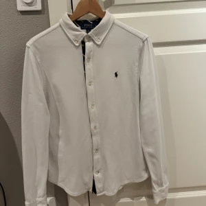 Vit skjorta  Polo Ralph Lauren - Vit skjorta från Polo Ralph Lauren med klassisk krage och knappar framtill. Liten broderad logga på bröstet och snygga detaljer i kragen med marinblått tyg, röda och vita ränder samt små gula hästar. Skjortan har lång ärm och är gjord i mjuk bomull. Sitter som en xxs!
