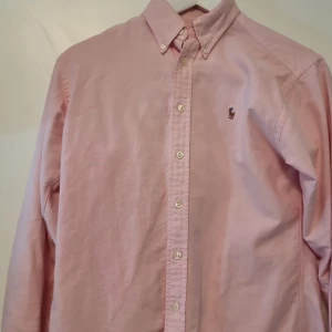 Rosa skjorta från Ralph Lauren - Snygg rosa skjorta från Ralph Lauren med klassisk button-down krage och broderad logga på bröstet. Skjortan har långa ärmar, vita knappar och är gjord i mjuk bomull. Perfekt för dig som gillar stilrena och tidlösa plagg.
