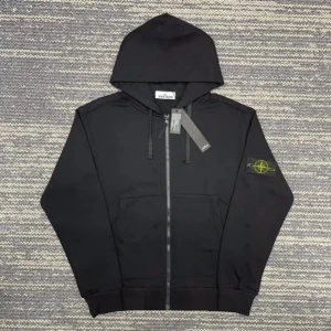 Svart hoodie från Stone Island - Svart hoodie från Stone Island med dragkedja och huva. Klassisk logotyp-patch på vänster ärm i gult och grönt. Tröjan har långa ärmar, ribbade muddar och snörning vid huvan. Tillverkad i mjuk bomull, perfekt för chill och streetwear.