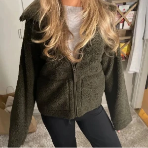Mörkgrön teddyjacka med dragkedja - Säljer en mörkgrön teddyjacka med mysigt fluffigt material och hel dragkedja framtill. Jackan har breda långa ärmar och en skön krage som ger extra stilpoäng. Perfekt för dig som gillar chill och comfy vibes.