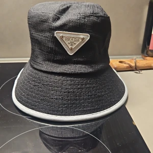 Svart bucket hat - Stilren svart bucket hat med vit kant och ikonisk metallogo framtill. Hatten har en lätt texturerad yta och klassisk form. Perfekt accessoar för att lyfta din look med exklusiv känsla.