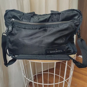 Väska Diesel -  Perfekta herrväskan från Diesel med flera fack och robusta dragkedjor. Väskan har både handtag och justerbar axelrem, samt tydlig Diesel-logga på båda sidor. Tillverkad i slitstark polyester med snygga skinn och metall-detaljer. toppendesign för vardagsbruk. 