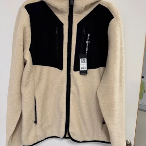 Sail Racing Patrol Pile Hood - XL Herr - Säljer en helt ny beige och svart teddyjacka från Sail Racing i storlek XL. Jackan har huva, hel dragkedja framtill och två svarta bröstfickor med dragkedja. Materialet är mjuk fleece och jackan har en clean, sportig look med broderad logga på bröstet. Storlek: XL. Nypris: 1999:-.