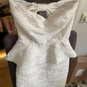 Vit kort klänning med rosett - Supersöt vit kort klänning med struktur i tyget och stor rosett i ryggen. Klänningen har en figurnära passform och är axelbandslös, perfekt för dig som vill sticka ut. Materialet känns lyxigt och har en snygg rynkad effekt.passar storlek 34-36 mycket elegant vit klänning. Mycket bra skick 