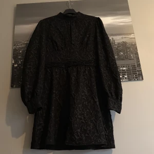 Svart kortklänning från SHEIN - Svart kortklänning från SHEIN med långärmade puffärmar och subtilt blommönster i tyget. Klänningen har hög hals, markerad midja med dekorativa ränder och dragkedja bak. Materialet är syntet och har en lätt glansig finish som ger en cool vibe.