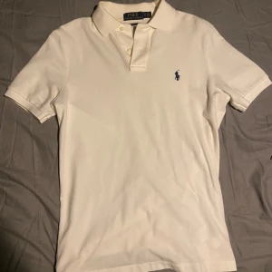 Vit pikétröja från Ralph Lauren - Vit pikétröja från Ralph Lauren med klassisk krage och korta ärmar. Tröjan har blå broderad logga på bröstet och knappar vid halsen. Tillverkad i mjuk bomull som känns skön mot huden. Perfekt för en clean och stilren look.