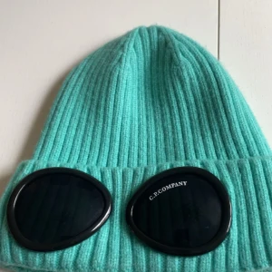 Turkos ribbad goggle-mössa C.P. Company - Säljer en turkos ribbstickad mössa från C.P. Company med ikoniska svarta goggle-linser framtill. Mössan är tillverkad i mjuk ull och har en uppvikt kant med märkets logga på ena linsen. Perfekt för dig som vill sticka ut med streetwear-vibe.