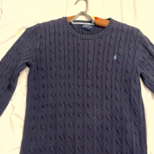 Mörkblå kabelstickad tröja Polo Ralph Lauren - Kabelstickad mörkblå tröja från Polo Ralph Lauren med rund halsringning och broderad logga på bröstet. Tröjan har lång ärm och klassisk passform, perfekt för lager-på-lager-stil. Tillverkad i mjuk bomull för en skön känsla.