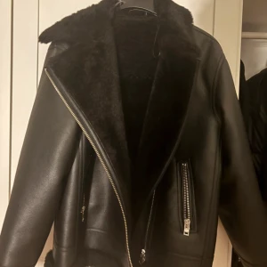 Svart teddyjacka med dragkedja - Snygg svart teddyjacka med bred krage och mjukt foder. Jackan har en cool asymmetrisk dragkedja framtill och två fickor med dragkedja. Materialet är en mix av skinn och teddy, vilket ger en lyxig och trendig vibe. Perfekt för dig som gillar streetstyle och vill hålla dig varm.