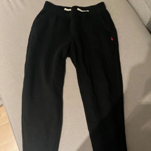 Svarta mjukisbyxor från Polo Ralph Lauren - Svarta mjukisbyxor från Polo Ralph Lauren med vit snörning i midjan och klassisk röd Polo-logga broderad vid fickan. Byxorna har normal passform och är gjorda i mjukt bomullsmaterial, perfekta för chill dagar eller träning.