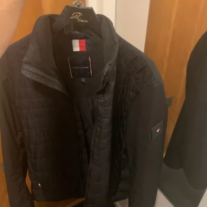 Svart quiltad jacka Tommy Hilfiger - Svart quiltad jacka från Tommy Hilfiger i storlek M. Jackan har dragkedja framtill, diskret Hilfiger-logga på bröstet och ärmen samt ribbade muddar. Materialet är syntet och polyester, perfekt för höst och vår. Snygg och stilren design med normal passform.