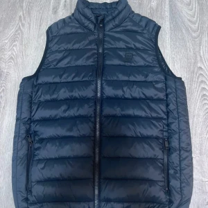 Svart dunväst från Jack & Jones - Snygg svart dunväst från Jack & Jones med quiltad design och hög krage. Västen har två praktiska fickor med dragkedja och en diskret logotyp på bröstet. Perfekt att slänga över en hoodie för extra värme under kyliga dagar.