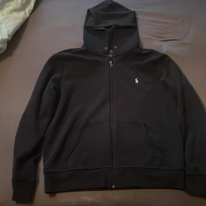 Mörk blå hoodie från Polo Ralph Lauren - Mörk blå hoodie från Polo Ralph Lauren med klassisk broderad logga på bröstet. Tröjan har dragkedja framtill, huva med snörning och två fickor. Perfekt för en chill och stilren look.