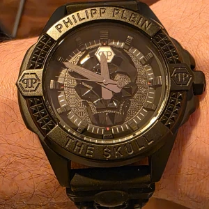 Philipp Plein Titan Black klockan  - Riktigt fet svart klocka från Philipp Plein med cool döskalle på urtavlan och armbandet. Klockan har detaljerade nitar runt boetten och tydlig logga. Armbandet är i matt svart material och hela looken är mörk och edgy. Perfekt för dig som gillar statement-accessoarer.