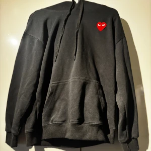 Svart hoodie Comme des Garçons Play - Svart hoodie från Comme des Garçons Play med klassisk röd hjärtlogga på bröstet. Tröjan har huva med snörning, känguruficka och ribbade muddar. Perfekt för dig som gillar streetstyle och vill ha något stilrent men ändå med en twist. Passar både storlek M och S. Använd Max 3 gånger.