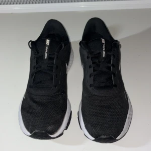 Nike Revolution 5 svarta sneakers - Nike Revolution 5 sneakers i svart med vit sula och vit swoosh på sidan. Skorna har mesh-överdel för bra ventilation, snörning och platt sula med dämpning. Perfekta för dig som gillar sportig stil och vill ha bekväma skor till träning eller vardag.