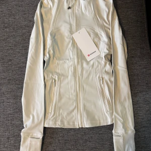 Lululemon define jacka - Snygg vit träningsjacka från Lululemon med figurnära passform och långa ärmar. Jackan har dragkedja framtill, två fickor med dragkedja och coola sömdetaljer. Materialet är mjukt och stretchigt, perfekt för aktiva dagar. Lululemon-logga på ryggen.