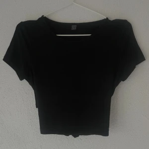 Svart croppad topp med knyt från SHEIN - Svart croppad topp från SHEIN i storlek S. Toppen har korta ärmar och rund halsringning. Baksidan har en öppen detalj med långa band som kan knytas för en snygg look. Mjuk och stretchig, perfekt till jeans eller kjol.