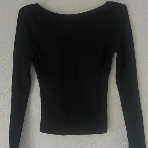 Svart ribbad långärmad topp - Säljer en svart långärmad topp med ribbad struktur och djup rundad halsringning. Modellen är slim och croppad, vilket ger en snygg siluett. Materialet är mjukt och stretchigt, perfekt för att styla med jeans eller kjol. Enkel och stilren design.
