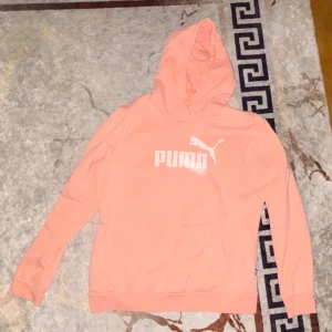 Ljusrosa hoodie från Puma - Säljer en ljusrosa hoodie från Puma med vit logga på bröstet. Tröjan har huva, lång ärm och en stor magficka framtill. Perfekt för dig som gillar sportiga och bekväma plagg med stilren design.