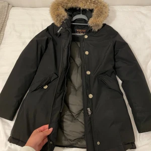 Svart parkas från Woolrich med päls M nyskick  - Parkas från Woolrich i svart med beige päls på huvan. Jackan har långa ärmar, dragkedja och knappar framtill samt två fickor med lock. Insidan är quiltad för extra värme och huvan är fodrad med fluffig päls. Perfekt för kalla dagar. Endast några gånger använd! Nyskick, säljer för den har blivit liten på mig. 