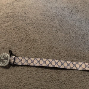 Guci bälte - Säljer ett snygg bälte med klassiskt GG-mönster i blått på beige bakgrund. Bältet har ett stort, rundat spänne i silve. Perfekt accessoar för att lyfta din outfit och visa din stil.