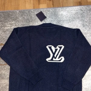 Mörkblå stickad Louis Vuitton tröja - Exklusiv mörkblå stickad tröja från Louis Vuitton med stor vit LV-logga på bröstet. Rund halsringning och ribbade muddar vid ärmslut och nederkant. Perfekt för dig som vill ha en lyxig och stilren look med designer vibe.