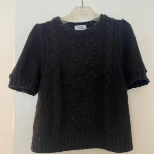 Svart stickad tröja med kort ärm - Svart stickad tröja från A Better Button med korta ärmar och snygga kabelstickade detaljer framtill. Tröjan har ribbade muddar vid ärmslut och nederkant samt ett coolt strukturmönster med små bollar. Perfekt för dig som gillar en chill och trendig look.