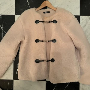 Beige teddyjacka från Gina Tricot - Säljer en beige teddyjacka från Gina Tricot med coola svarta detaljer och metallspännen framtill. Jackan har rund hals, lång ärm och är gjord i mjuk fleece som ger en fluffig vibe. Perfekt för dig som gillar en chill och trendig look.