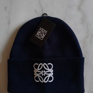 Mörkblå Loewe mössa med broderi - Snygg mörkblå mössa från Loewe med vit broderad logga framtill. Klassisk uppvikt kant och mjukt stickat material i akryl. Perfekt för dig som vill ha en stilren och exklusiv look när det är kallt ute.