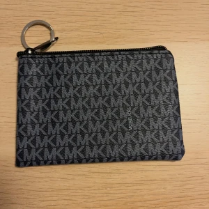 Svart plånbok från Michael Kors - Snygg plånbok från Michael Kors i svart med grått MK-mönster över hela ytan. Plånboken har dragkedja upptill och en metallring för nycklar eller att fästa i väskan. Perfekt storlek för kort och småsaker.