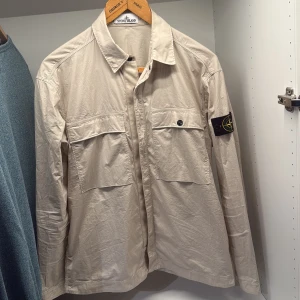 Beige overshirt från Stone Island - Snygg beige overshirt från Stone Island med klassisk krage och två stora bröstfickor med knappar. Ikonisk patch på vänster ärm. Tillverkad i lätt bomullsmaterial, perfekt för lager-på-lager. Passar dig som gillar clean och stilren streetwear.  Helt ny använd endast en gång. Storlek S men passar både S och M Nypris strack under 6k köpt på Nk