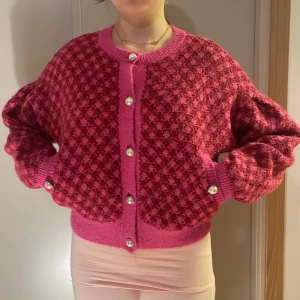 Rosa kofta med pärlknappar från Zara - Superfin rosa kofta från Zara med coolt rutigt mönster och stora pärlknappar. Koftan är stickad, har lång ärm och breda muddar. Perfekt för dig som vill sticka ut med en färgglad och trendig look.