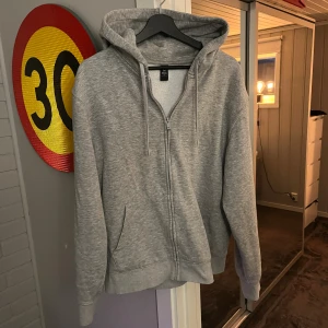 Grå hoodie med dragkedja från soc  - En enkel och stilren grå hoodie från soc med dragkedja framtill och två fickor. Tröjan har huva med snörning och är tillverkad i mjuk bomullsblandning. Perfekt för chill dagar eller när du vill ha något bekvämt på dig. 