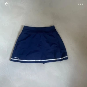 Mörkblå tenniskjol från Artengo - Sportig mörkblå tenniskjol från Artengo med inbyggda shorts under och vit rand längs fållen. Kjolen har en bekväm midja och är perfekt för dig som gillar att röra på dig. Snygg och funktionell design som ger en fräsch look på tennisbanan.