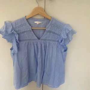 Ljusblå blus med volangärmar från Gina Tricot  - Supersöt ljusblå blus från Gina Tricot med v-ringning och volangärmar. Blusen har broderade detaljer och smock upptill, vilket ger en luftig och romantisk känsla. Perfekt för dig som gillar en feminin och chill stil. Materialet är mjuk bomull.