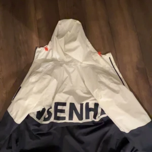 Vit och marinblå vindjacka Helly Hansen - Snygg vindjacka från Helly Hansen i storlek S. Jackan är vit upptill och marinblå nedtill med stor logga på ryggen. Den har huva och orange detaljer vid snörningen. Perfekt för blåsiga dagar och outdoor-aktiviteter.