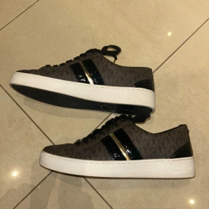  Michael Kors sneakers  - Snygga bruna sneakers från Michael Kors med MK-logga och mönstrad ovandel. Skorna har svarta och guldiga detaljer på sidan, svarta snören med guldfärgade ändar och vit platt sula. Insidan har guldfärgad logotyp och hälen är förstärkt med svart lackdetalj. OBS storleken är 7 vilket motsvarar ungefär 37-37,5 i storlek.