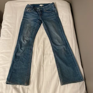 Blå bootcut jeans från Gina Tricot - Snygga blå jeans från Gina Tricot i storlek 158 med klassisk bootcut passform. Jeansen har fem fickor, kontrastsömmar och detaljerade bakfickor med lock och knapp. Perfekt för dig som gillar en avslappnad stil. Man kan ställa in midjan på jeansen.