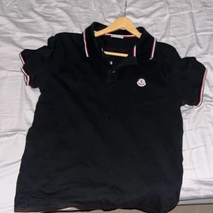 Svart Moncler pikétröja med röd/vit kant - Svart pikétröja från Moncler med klassisk krage och knappar framtill. Röd och vit kant på krage och ärmslut ger en snygg kontrast. Moncler-logga på bröstet. Perfekt för dig som gillar stilrena och sportiga plagg.