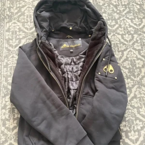 Moose knuckles bunny - Svart pufferjacka från Moose Knuckles i storlek S. Jackan har huva med snörning, guldfärgade detaljer och dragkedja framtill. Insidan är quiltad med mönstrat foder och jackan har en snygg logotyp på ärmen. Perfekt för kalla dagar och har en modern, urban vibe. Inga defekter köpt för va 6 tusen, kom privat för mer frågor! 🫎